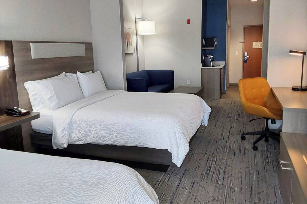A hotel room with two beds, a desk, a chair, a lamp, and a small kitchenette.