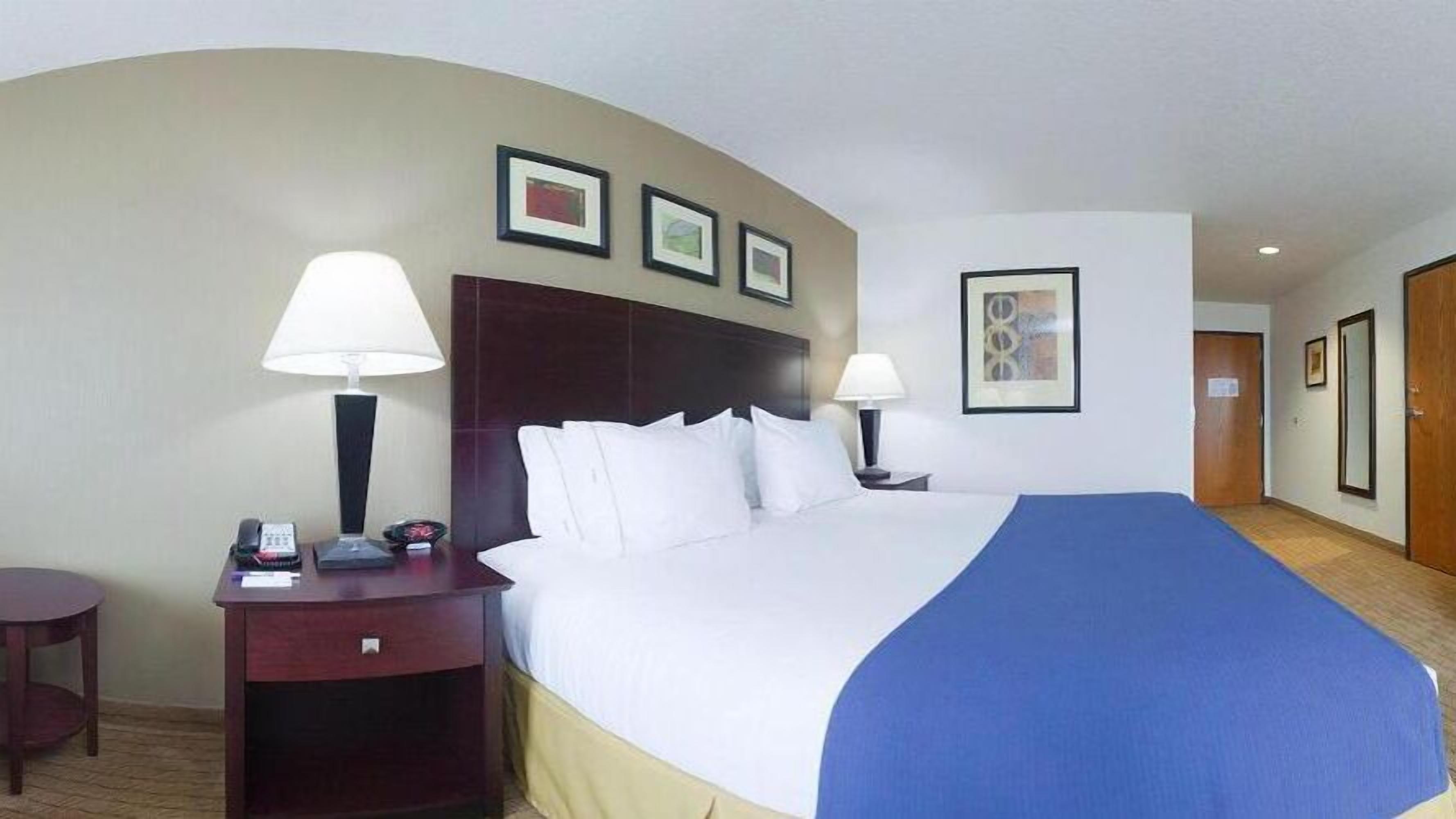 Una habitación de hotel con una cama grande, dos mesitas de noche, un escritorio y una silla. Se observan cuadros en la pared y una puerta a la derecha.