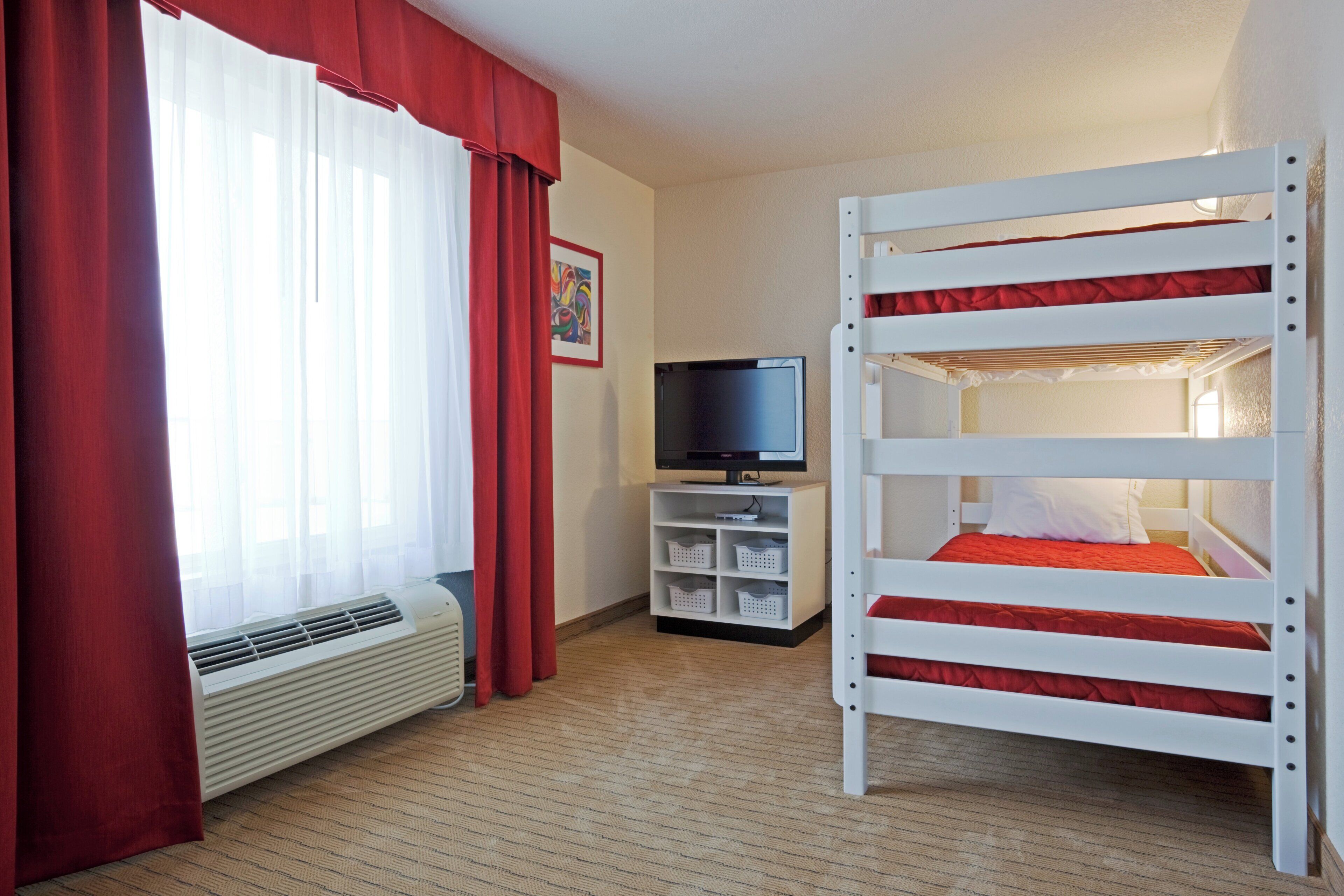 A hotel room with a bunk bed, a television, a small cabinet, and a window with curtains.