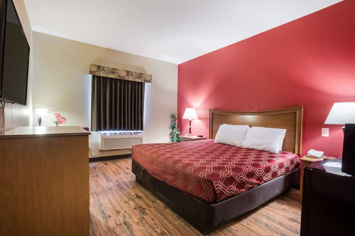 A hotel room with a large bed, a desk, a television, a window with curtains, and a red wall.