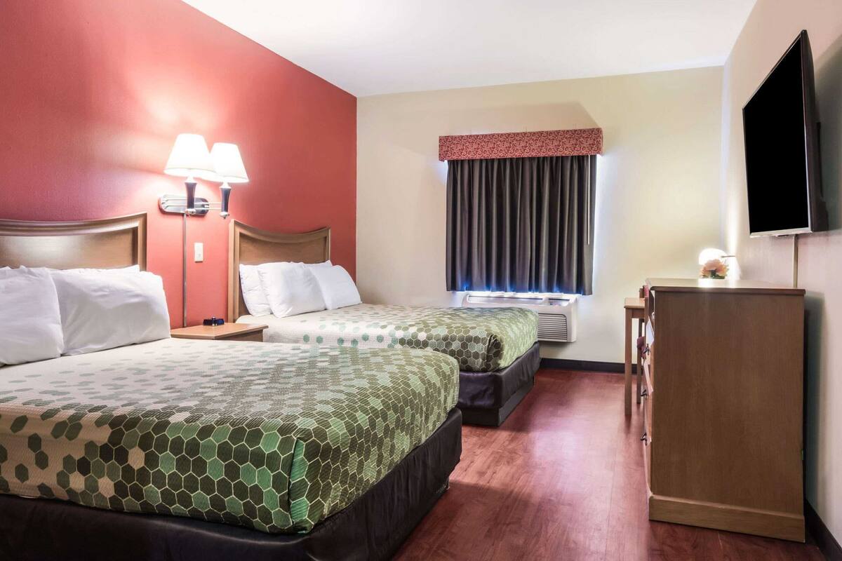 A hotel room with two beds, a flat-screen TV, a desk, and a window with curtains.