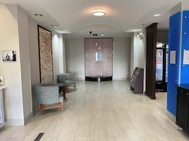 Uma área de recepção com chão de azulejos, duas poltronas, um quadro de informações fixado na parede e um balcão azul com um frigorífico.