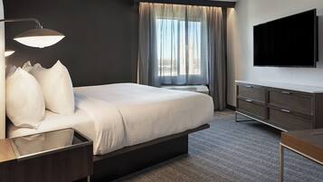 A hotel room with a bed, a desk, a TV, a window with curtains, and a herringbone-patterned carpet.