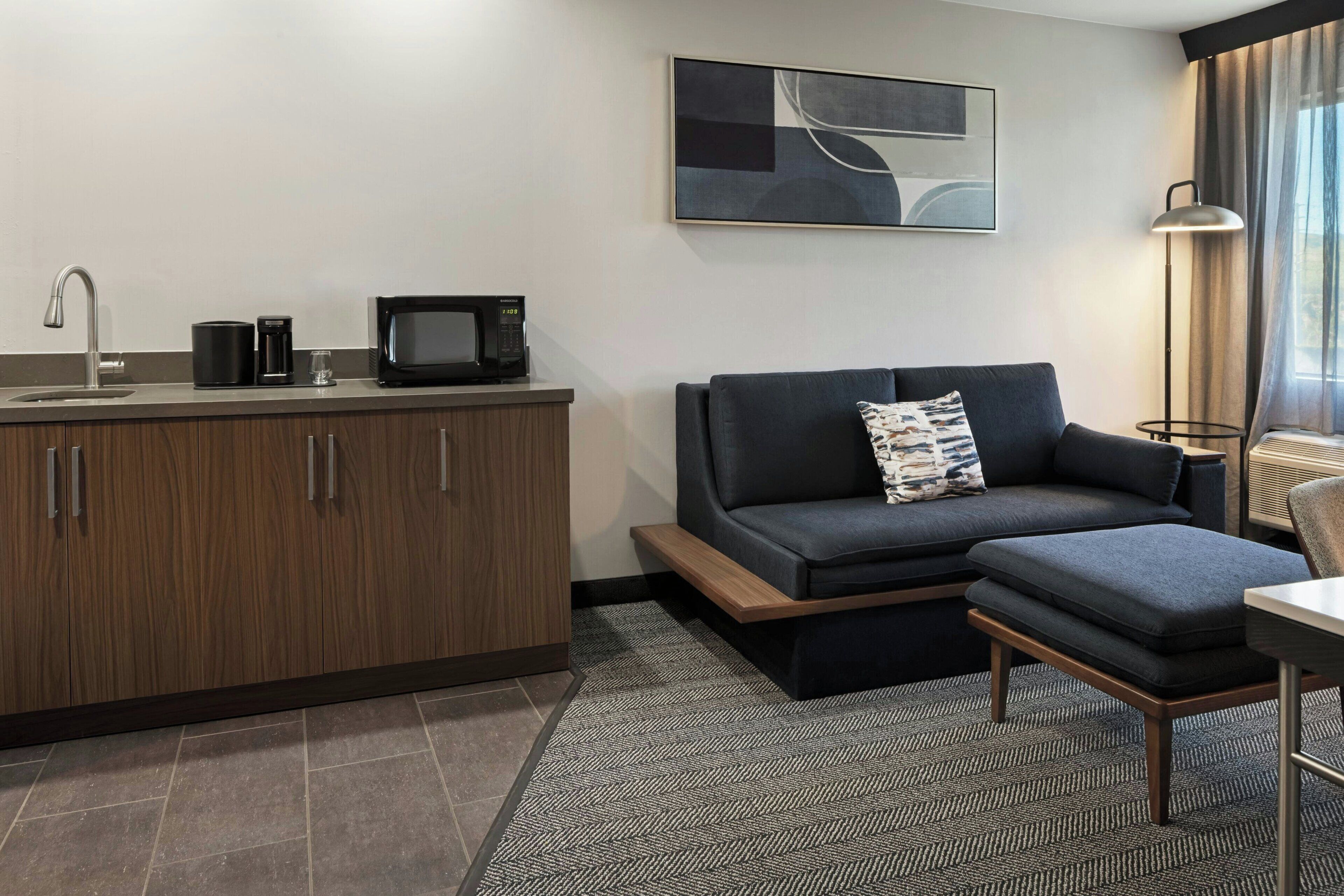 A modern hotel room with a kitchenette, a sofa, a small table, and a lamp.