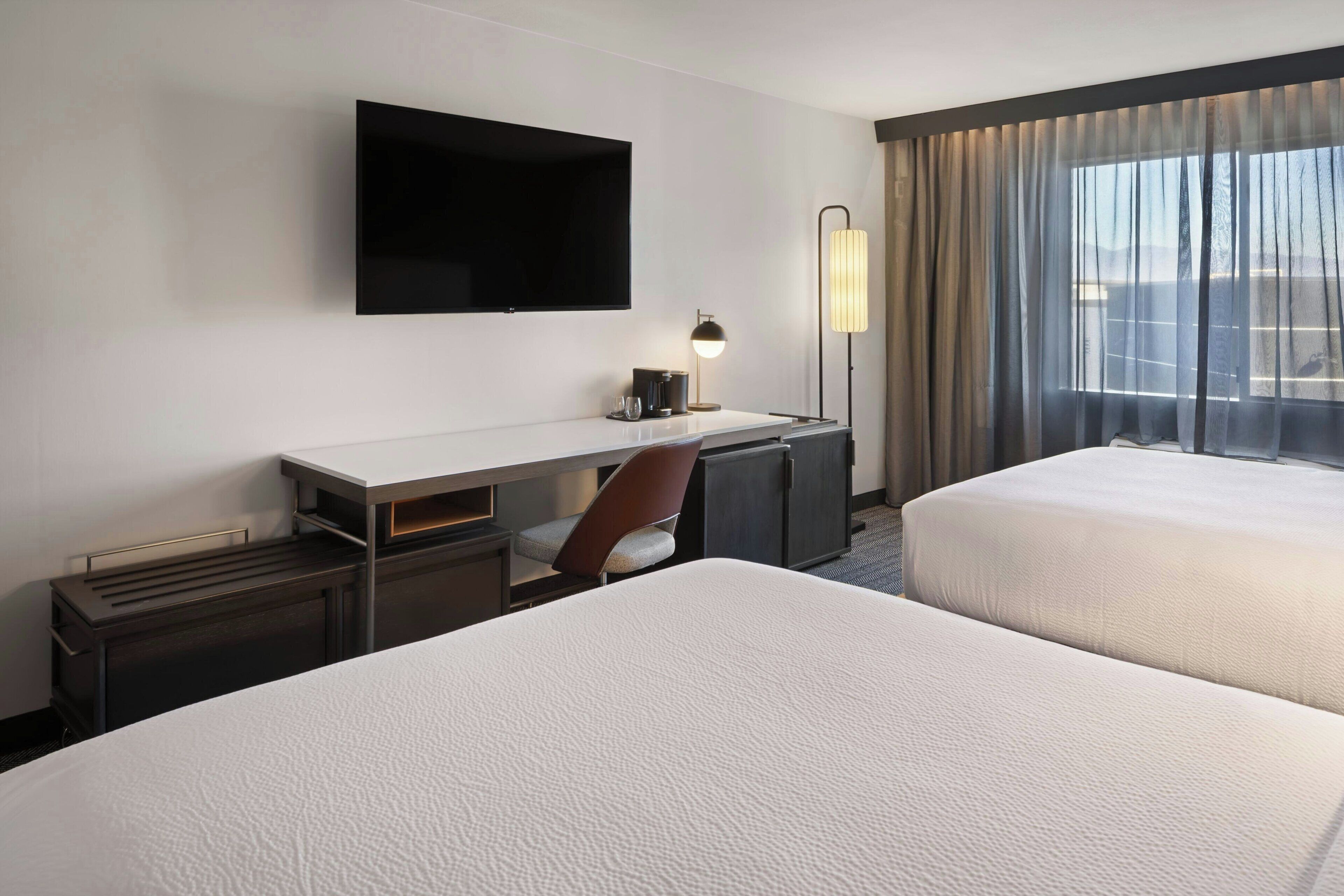 Ein Hotelzimmer mit zwei Betten, einem Schreibtisch mit Stuhl, einem Flachbildfernseher, einer Lampe und einem großen Fenster mit Vorhängen.