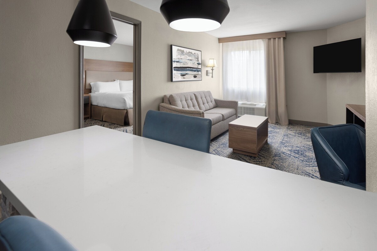Ein Hotelzimmer mit großem weißen Schreibtisch, einem beigen Sofa, einem blauen Sessel, einem kleinen Holztischchen, einem flachbildfernseher an der Wand und einem gerahmten Bild.