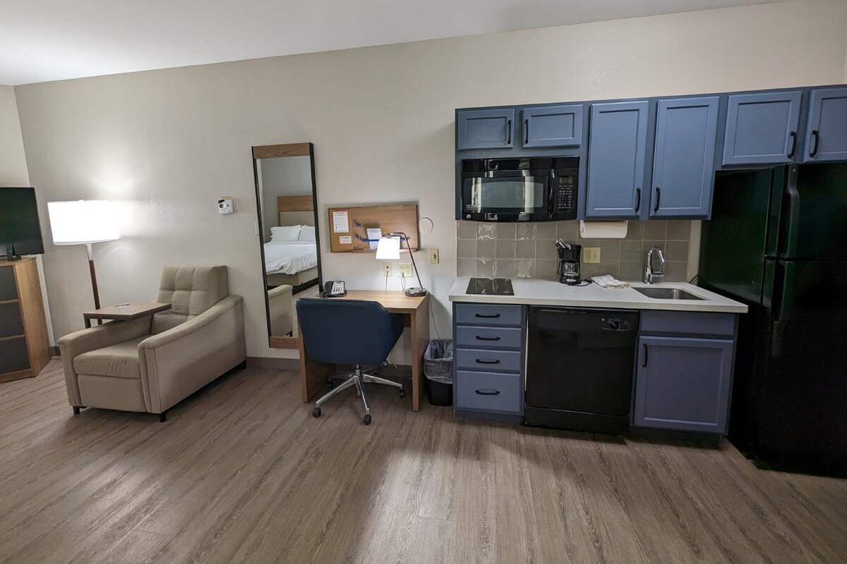 Ein kleines Hotelzimmer mit Küchenzeile, einem Bett, einem Schreibtisch und einem Stuhl.