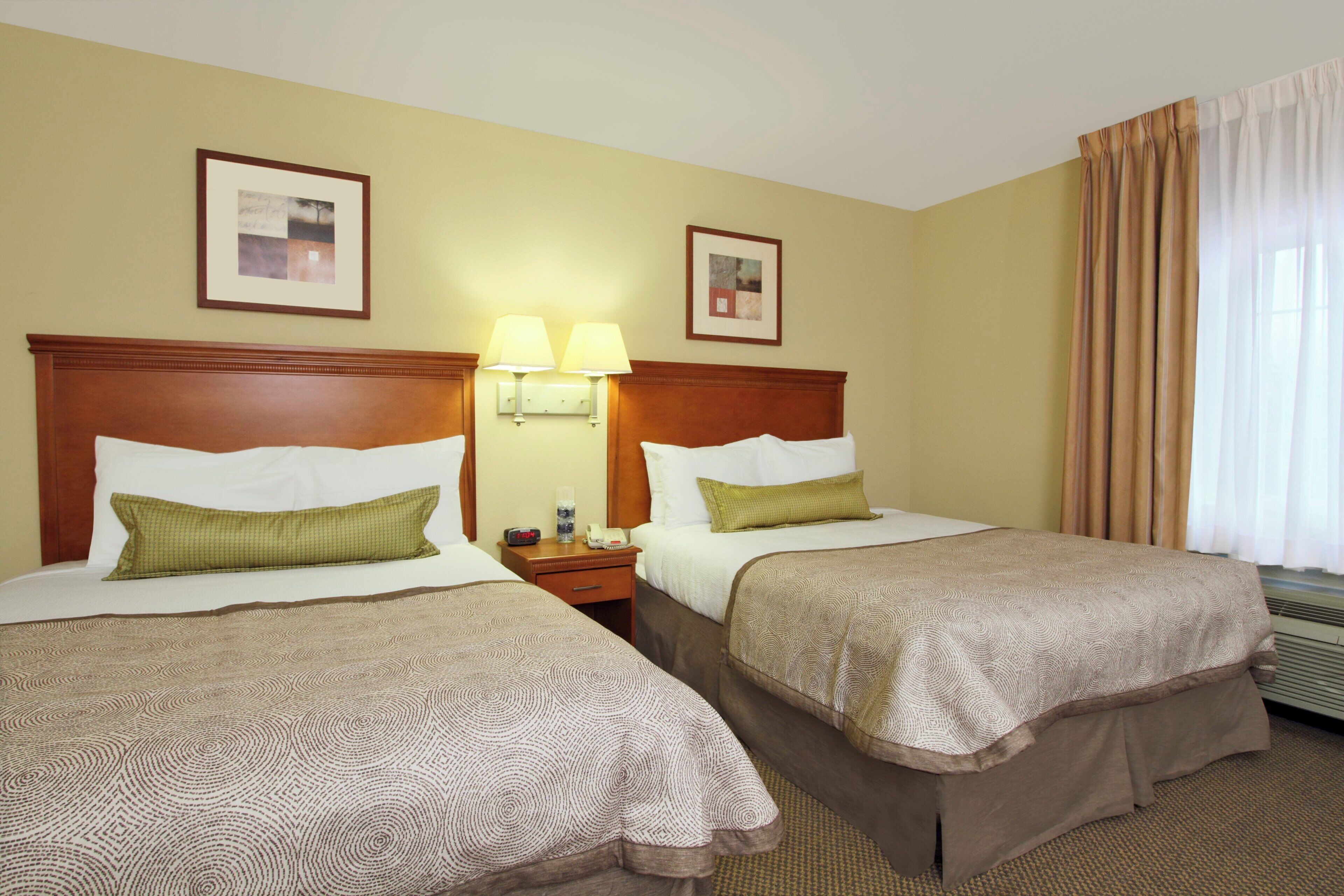 A hotel room with two beds, a nightstand, a lamp, and a window with curtains.