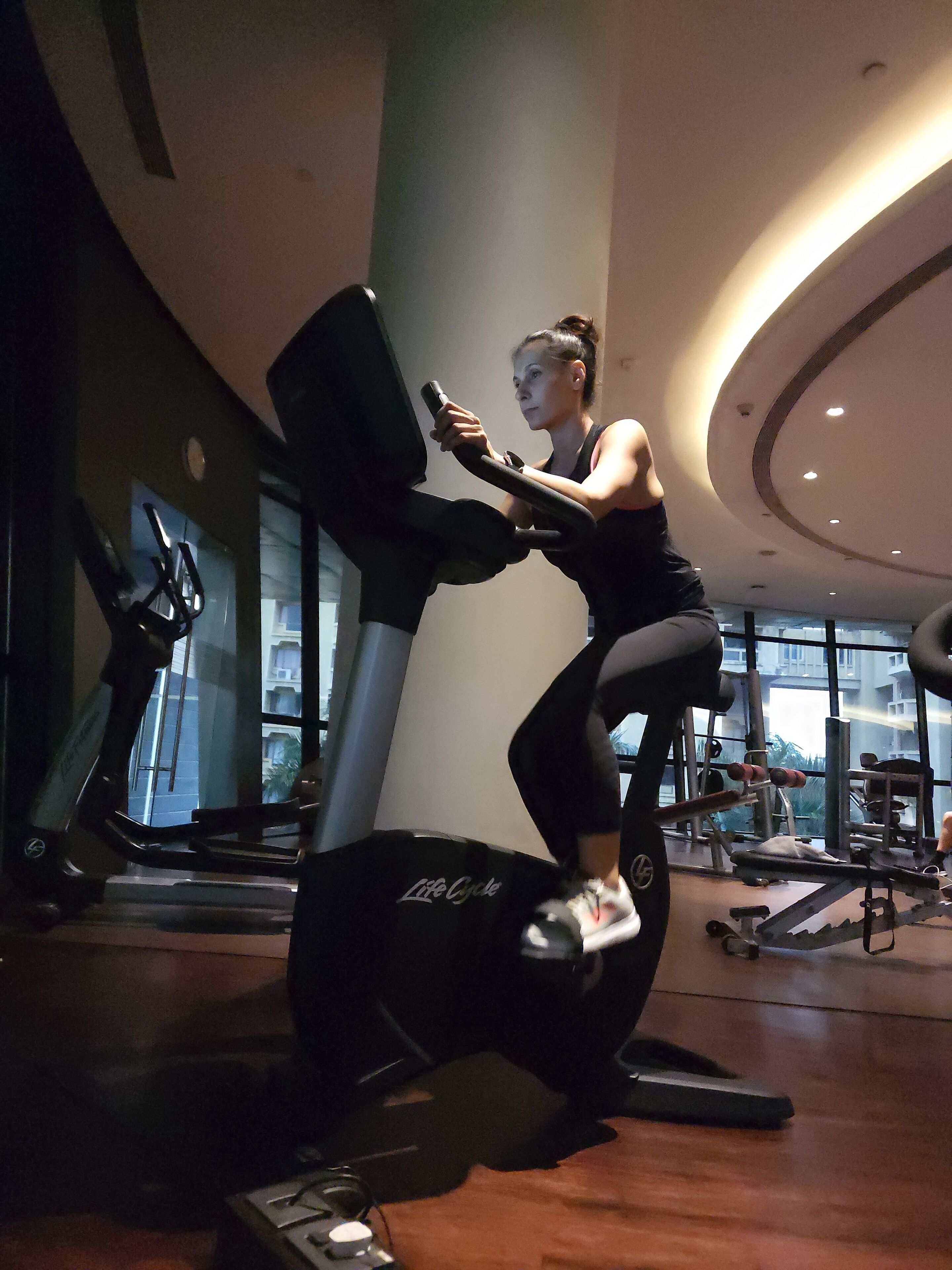 A woman using a stationary bike in a gym with various exercise equipment around.
