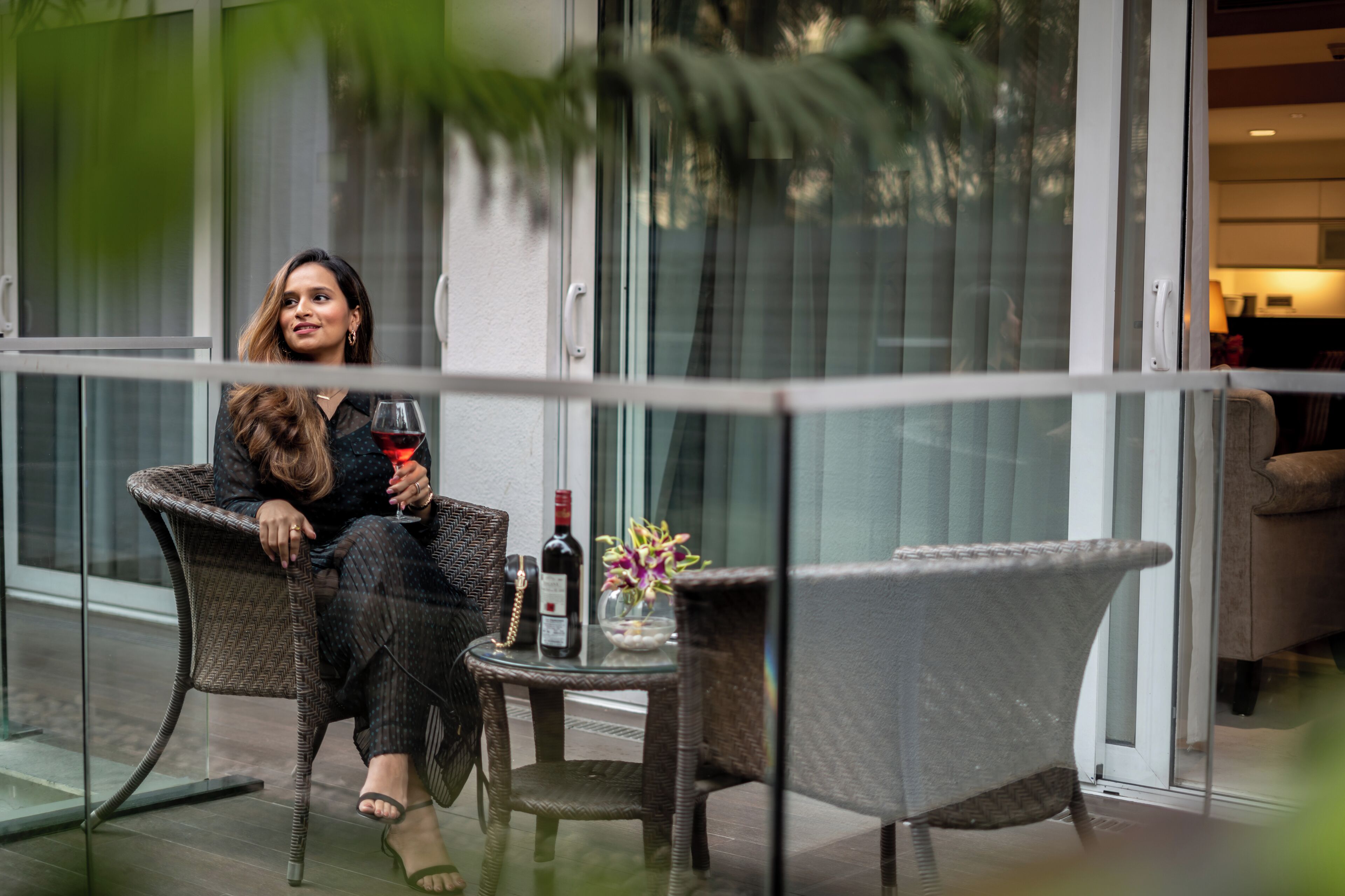 Una mujer sentada en una silla de mimbre en un balcón con una mesa de vidrio, sosteniendo una copa de vino tinto.