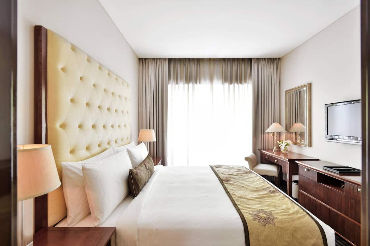 Ein Hotelzimmer mit hohem, rautenförmigem Kopfteil, einem Bett mit weißen Leinentextilien, einem Schreibtisch und einem Flachbildfernseher sowie einem großen Fenster mit Vorhängen.