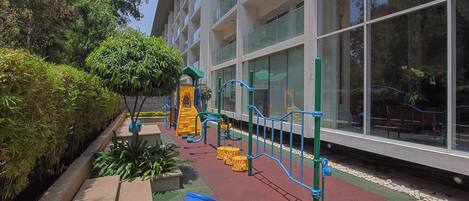 Un espace de jeux pour enfants avec un toboggan et des structures d’escalade, entouré d’un bâtiment doté de grandes fenêtres.
