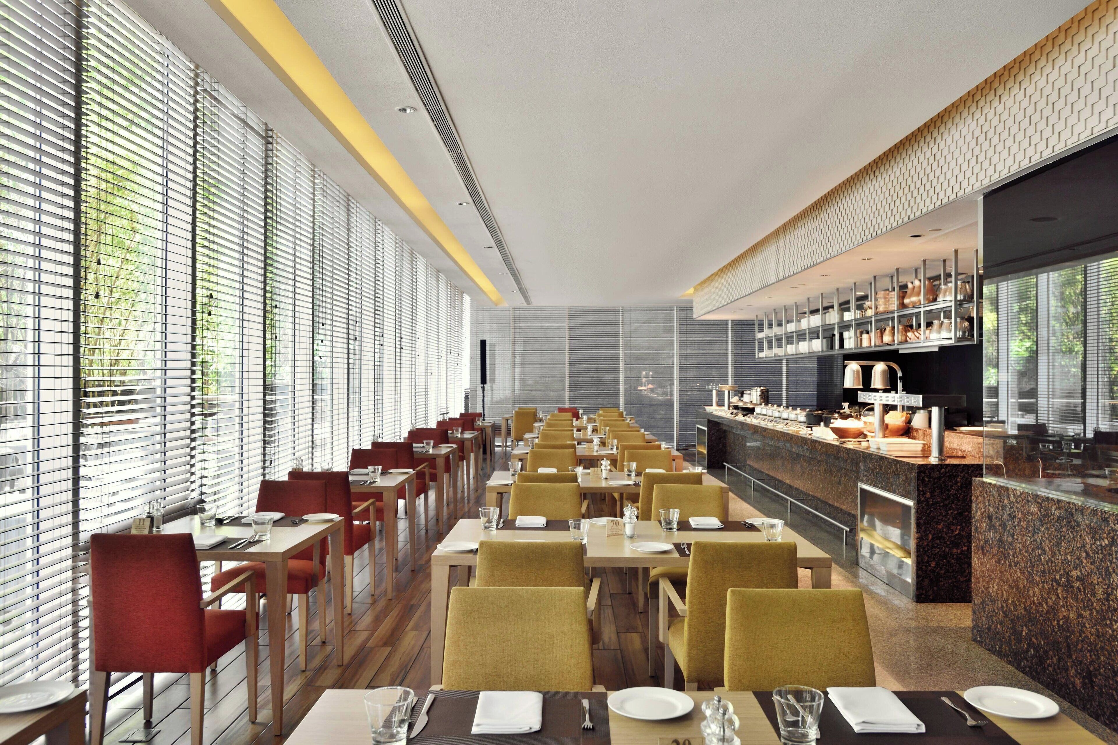 A modern restaurant with long tables and chairs, a counter with a display of food items, and large windows with blinds.
