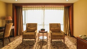 A hotel room with two armchairs, a small round table, a desk, and a lamp. The room has a large window with curtains and a view of the city. - Beijing Bao Lin Xuan International Hotel (Beijing)