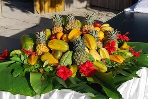 A table with a tropical fruit arrangement including pineapples, mangoes, and bananas, decorated with red flowers and green leaves. - The Islander Hotel (Praslin Island)