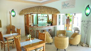 A dining area with wooden tables and chairs, a bar area with a thatched roof, and a view of the outside. - The Islander Hotel (Praslin Island)