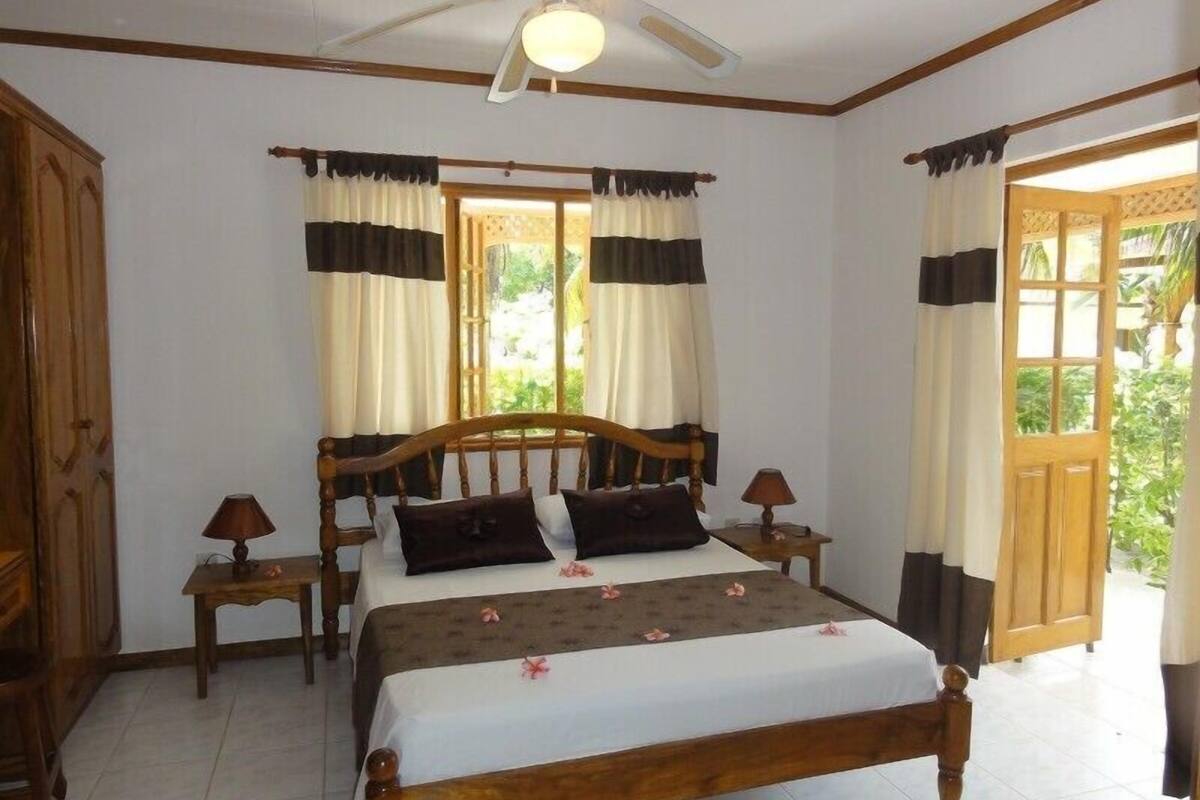 Quarto com cama de madeira, duas mesas de cabeceira, ventoinha no teto e janela com cortinas.