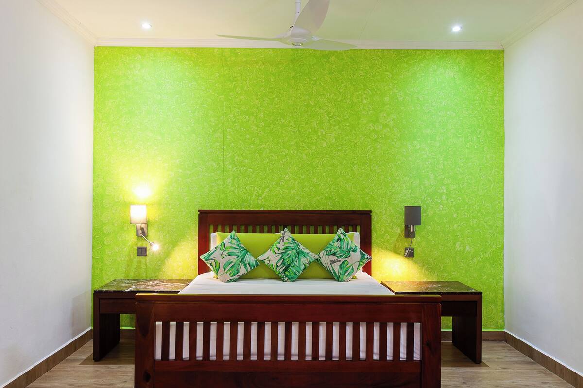 Quarto com parede estampada em verde, cama de madeira com duas almofadas, mesa de cabeceira e luminária na parede.