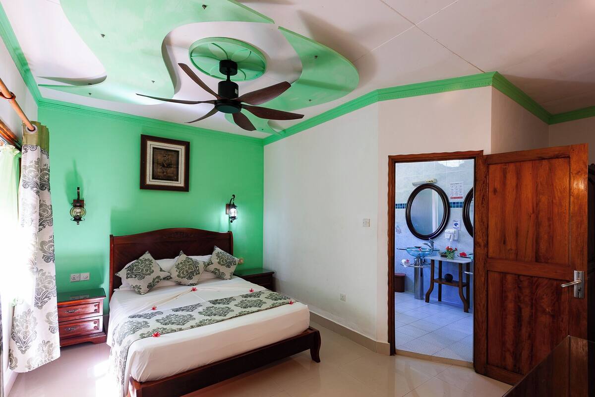 Quarto com cama, ventoinha no teto, mesa de cabeceira, cômoda, espelho e porta que dá para um banheiro.