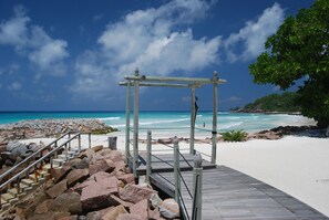 A wooden deck leading to a beach with a clear view of the ocean and a rocky shoreline. - The Islander Hotel (Praslin Island)