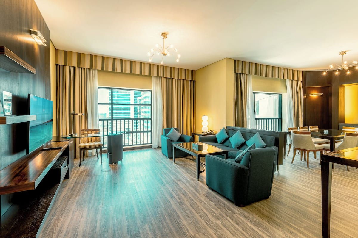 A modern hotel room with a large window, a sofa set, a dining table, and a TV.