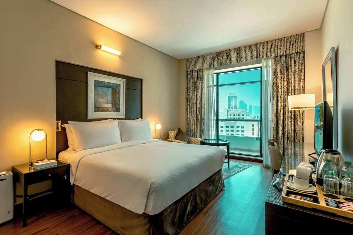 A hotel room with a large bed, bedside tables with lamps, a desk with a mirror, and a view of the city through the window.