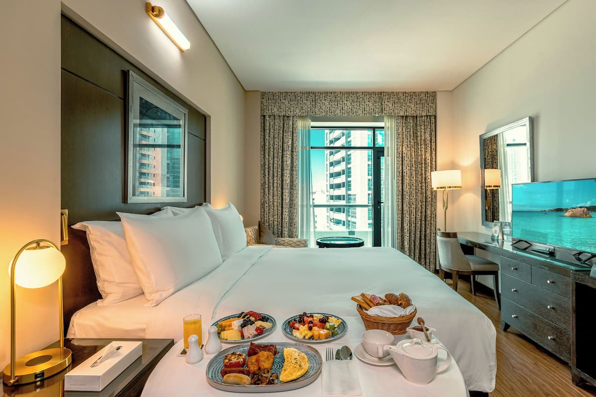 A hotel room with a bed, breakfast tray, bedside table, lamp, and a view of a building and water.