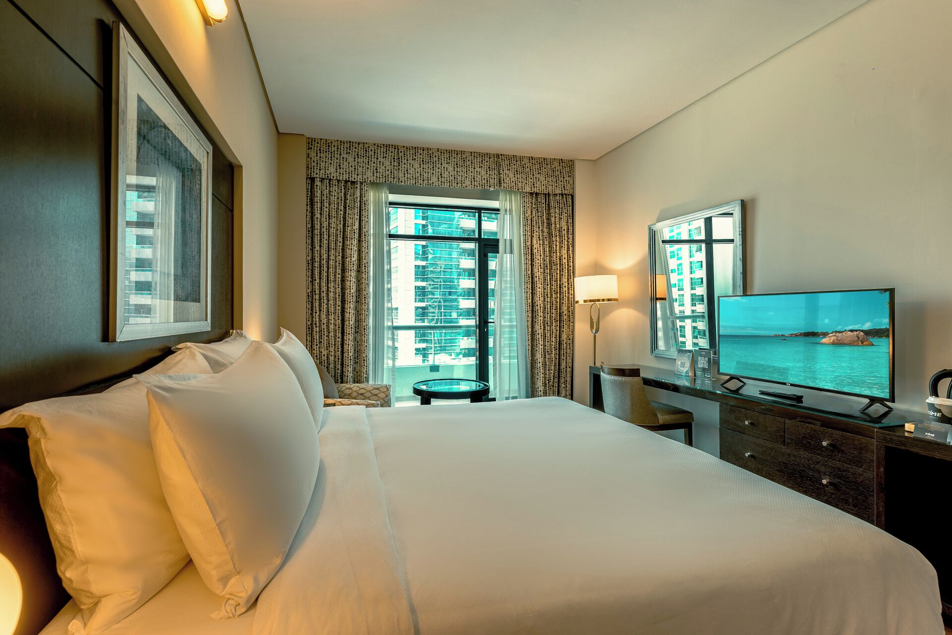 Habitación de hotel con una cama grande, un escritorio, televisor de pantalla plana, una silla y ventana con vistas a edificios y agua.