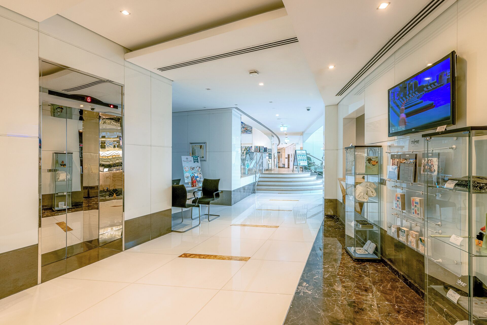 A modern hotel corridor with glass display cases, a staircase, and a television screen.