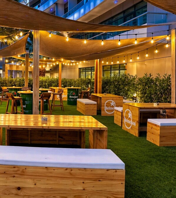 An outdoor seating area with wooden tables and chairs, branded with Heineken, under a canopy of string lights.