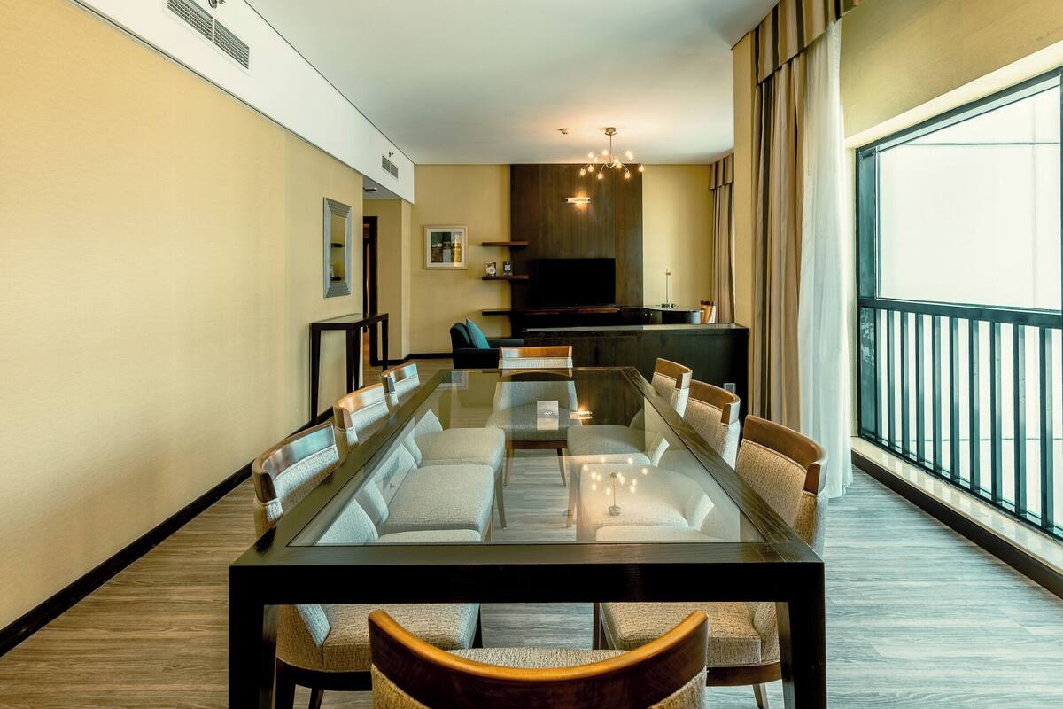 A modern dining area with a glass-top table and cushioned chairs, a television, and a balcony with a railing.