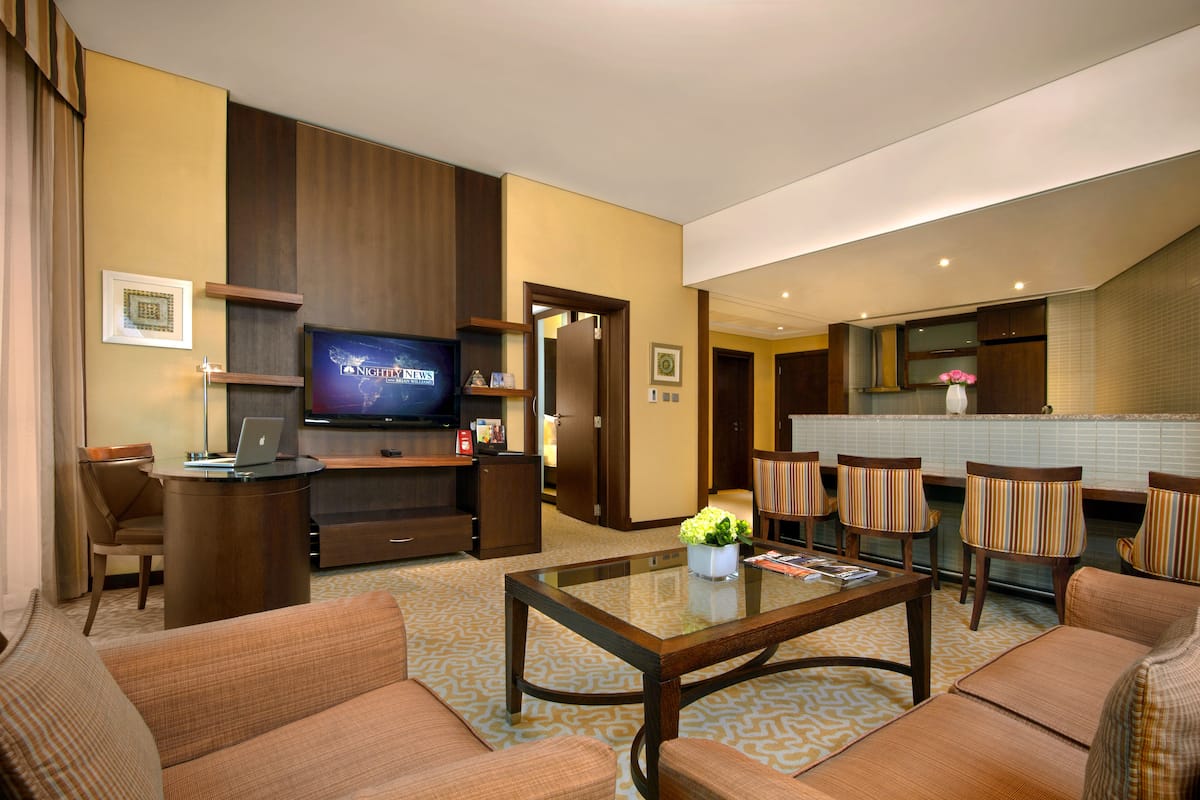 A hotel room with a living area, a television, a desk with a laptop, a dining area with striped chairs, and a kitchen with a white countertop.