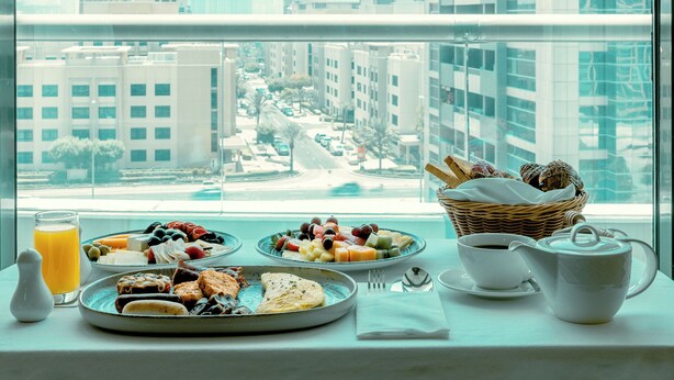 A breakfast setup with plates of food, a glass of orange juice, a basket of bread, and a teapot on a table by a window overlooking a cityscape. - TIME Oak Hotel & Suites (Dubai)