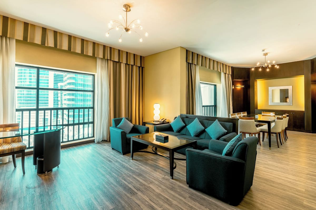 A modern hotel room with a large window, a sofa set, a dining table, and a chandelier.
