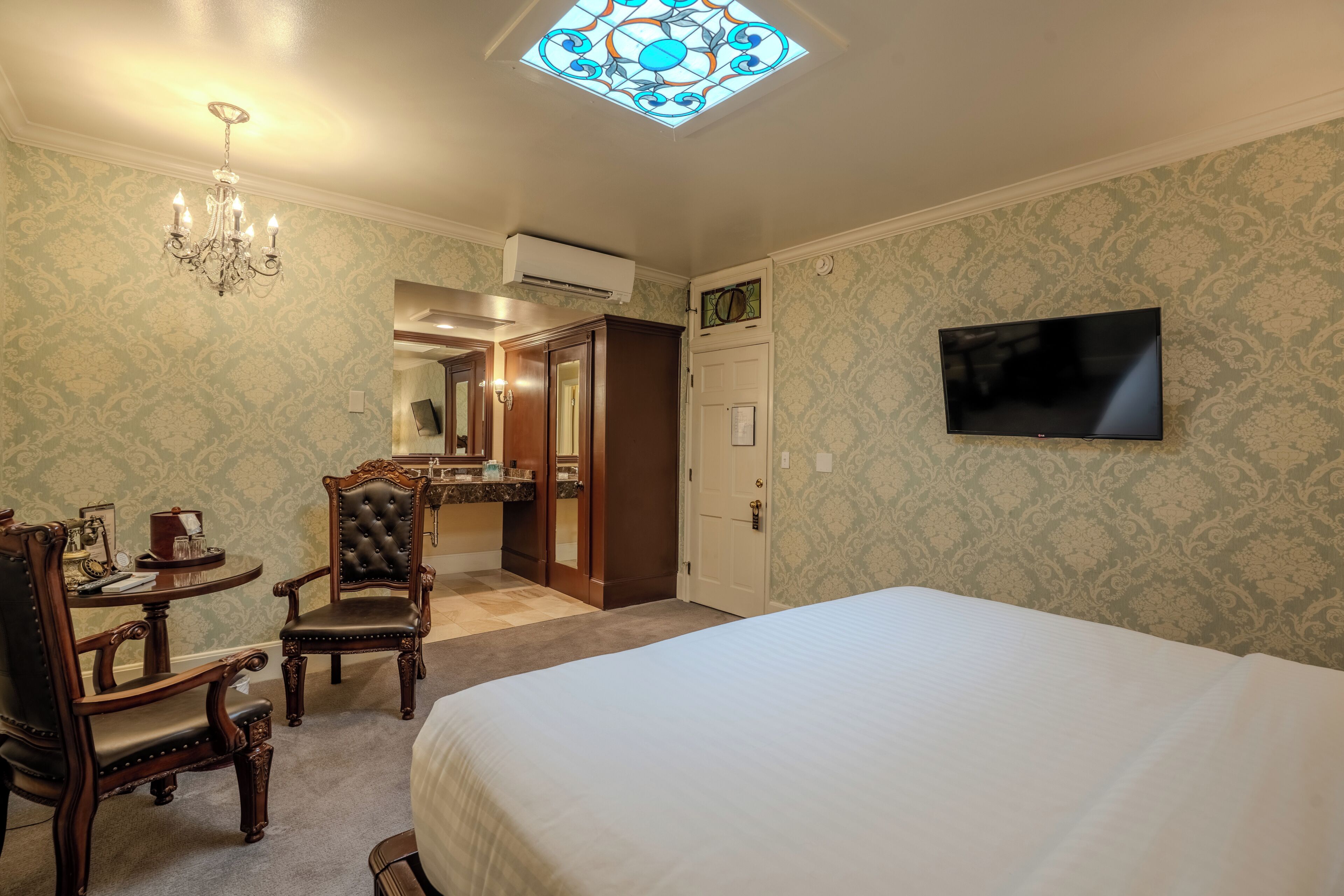 Habitación de hotel con cama, televisor, candelabro, escritorio con sillas y baño visible a través de una puerta abierta.
