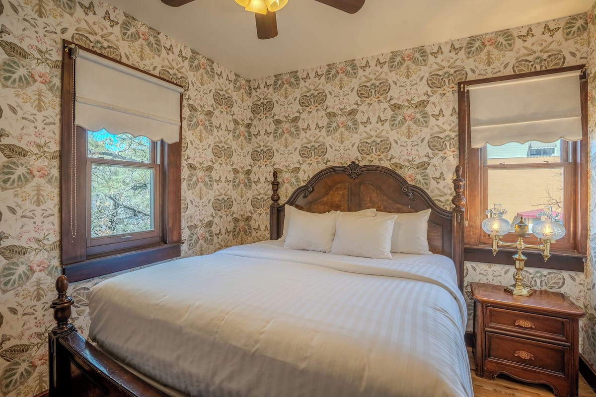 A bedroom with a wooden bed, floral wallpaper, a nightstand with a lamp, and a window with a view of trees.