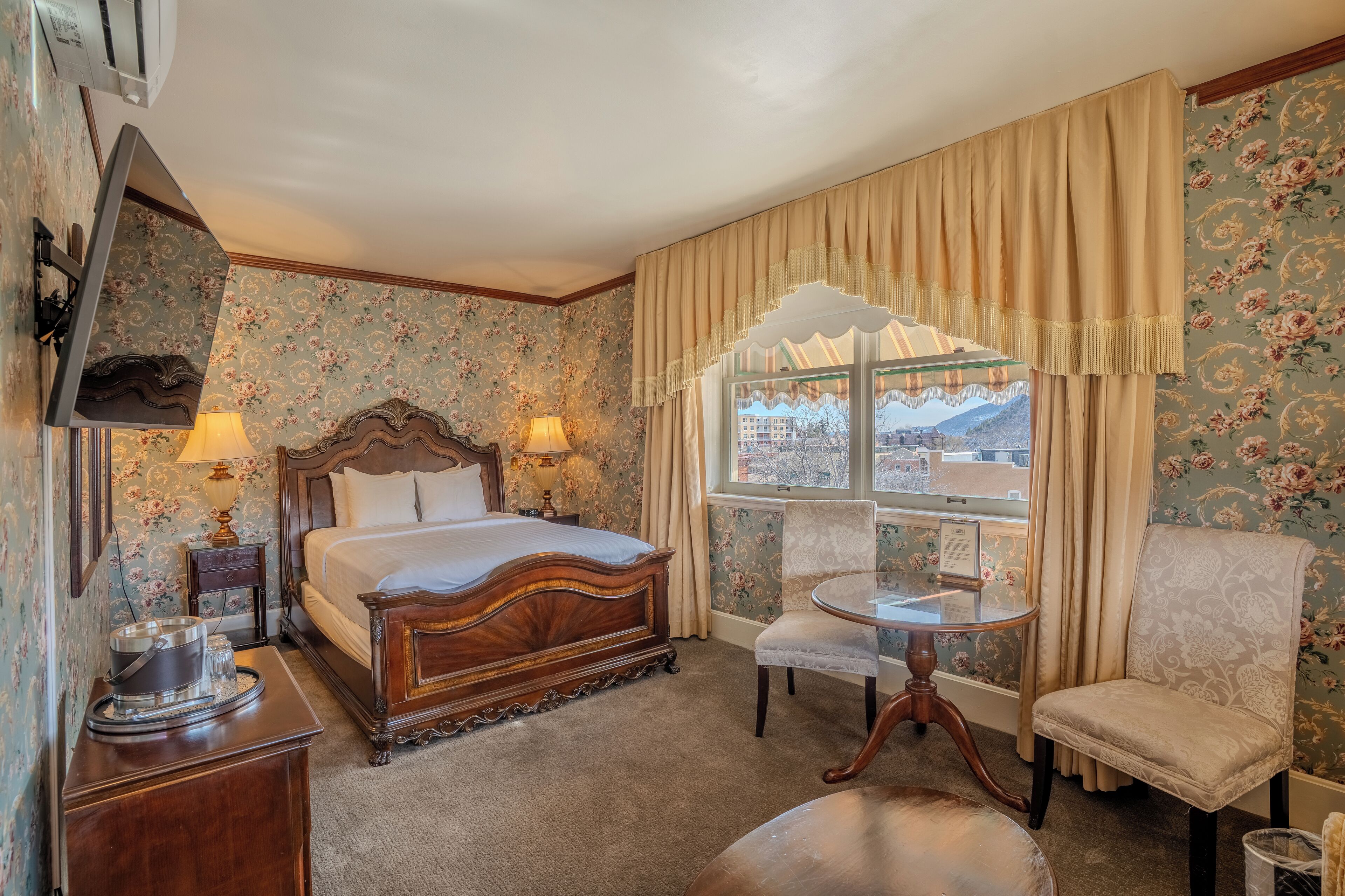 A classic hotel room with a large bed, a small table, a chair, and a window with curtains.