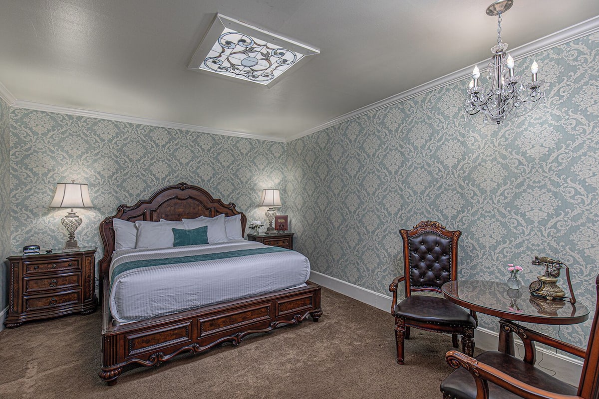A bedroom with a large bed, a wooden nightstand, a chair, a table with a lamp, and a chandelier.