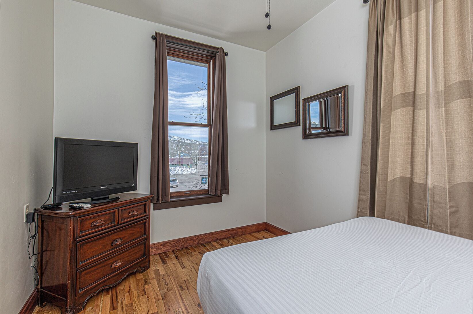A bedroom with a bed, a TV on a wooden dresser, a window with curtains, and two framed pictures on the wall.