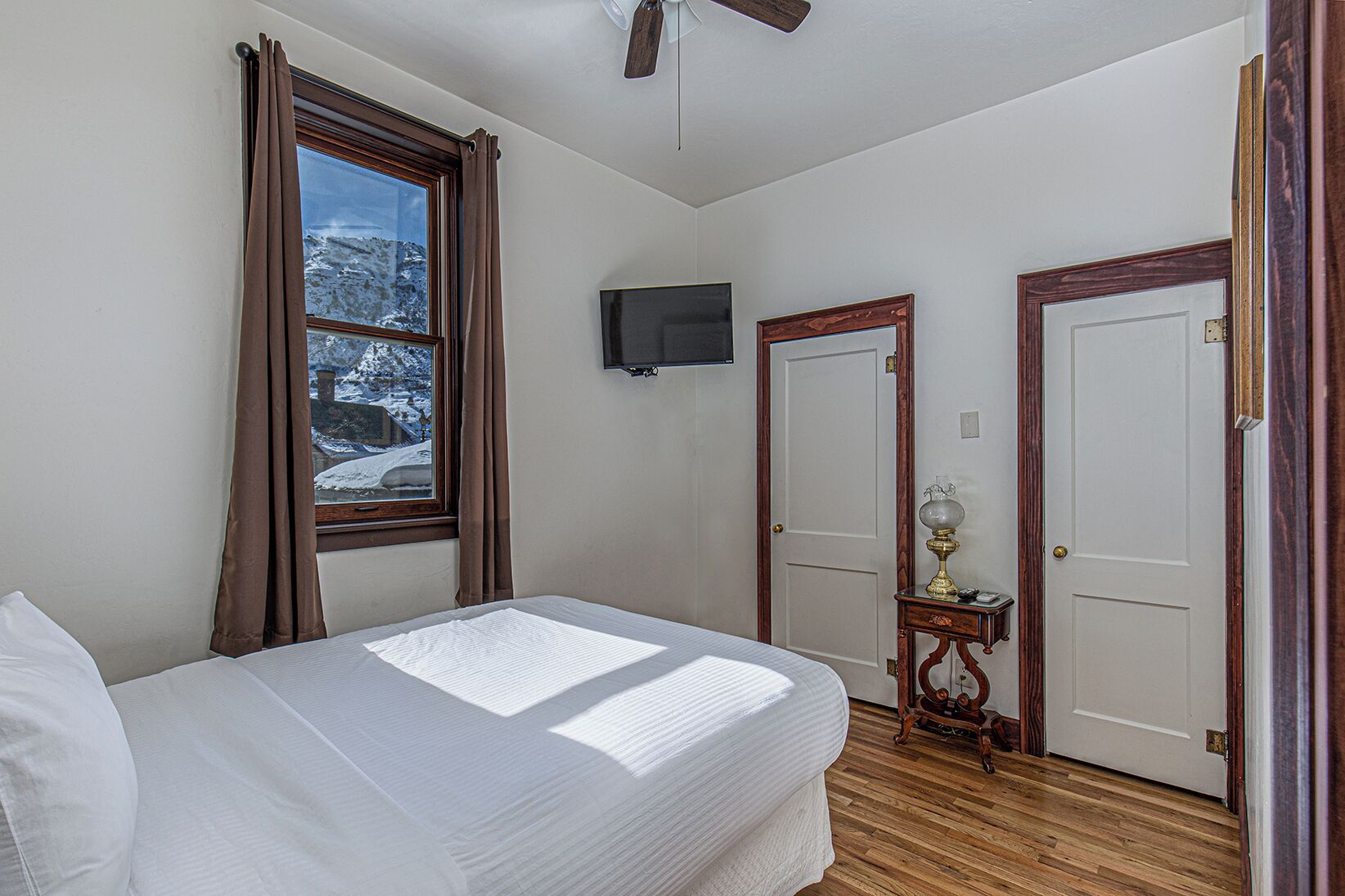 A bedroom with a bed, a ceiling fan, a TV mounted on the wall, and two doors.