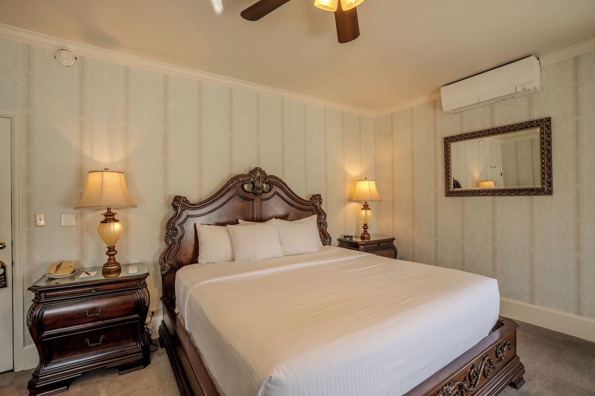 A neatly made bed in a hotel room with a wooden headboard, two bedside tables with lamps, a mirror, and an air conditioning unit on the wall.
