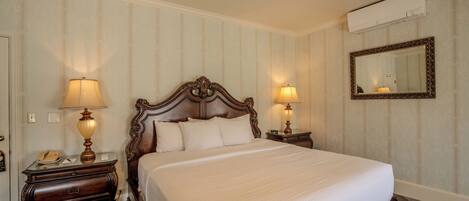 A neatly made bed in a hotel room with a wooden headboard, two bedside tables with lamps, a mirror, and an air conditioning unit on the wall.