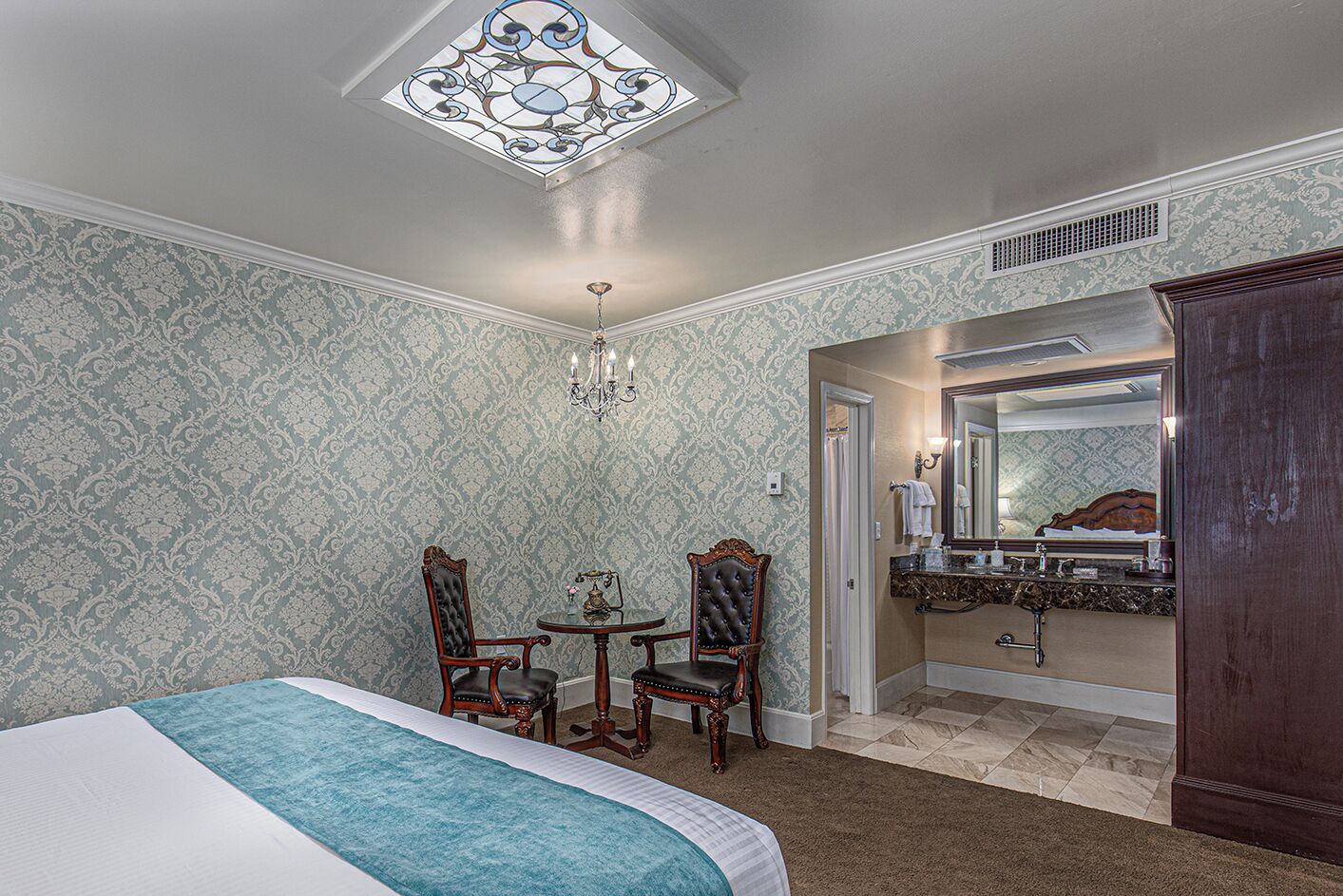 A bedroom with a bed, two chairs, a small table, a chandelier, a mirror, and a wardrobe.