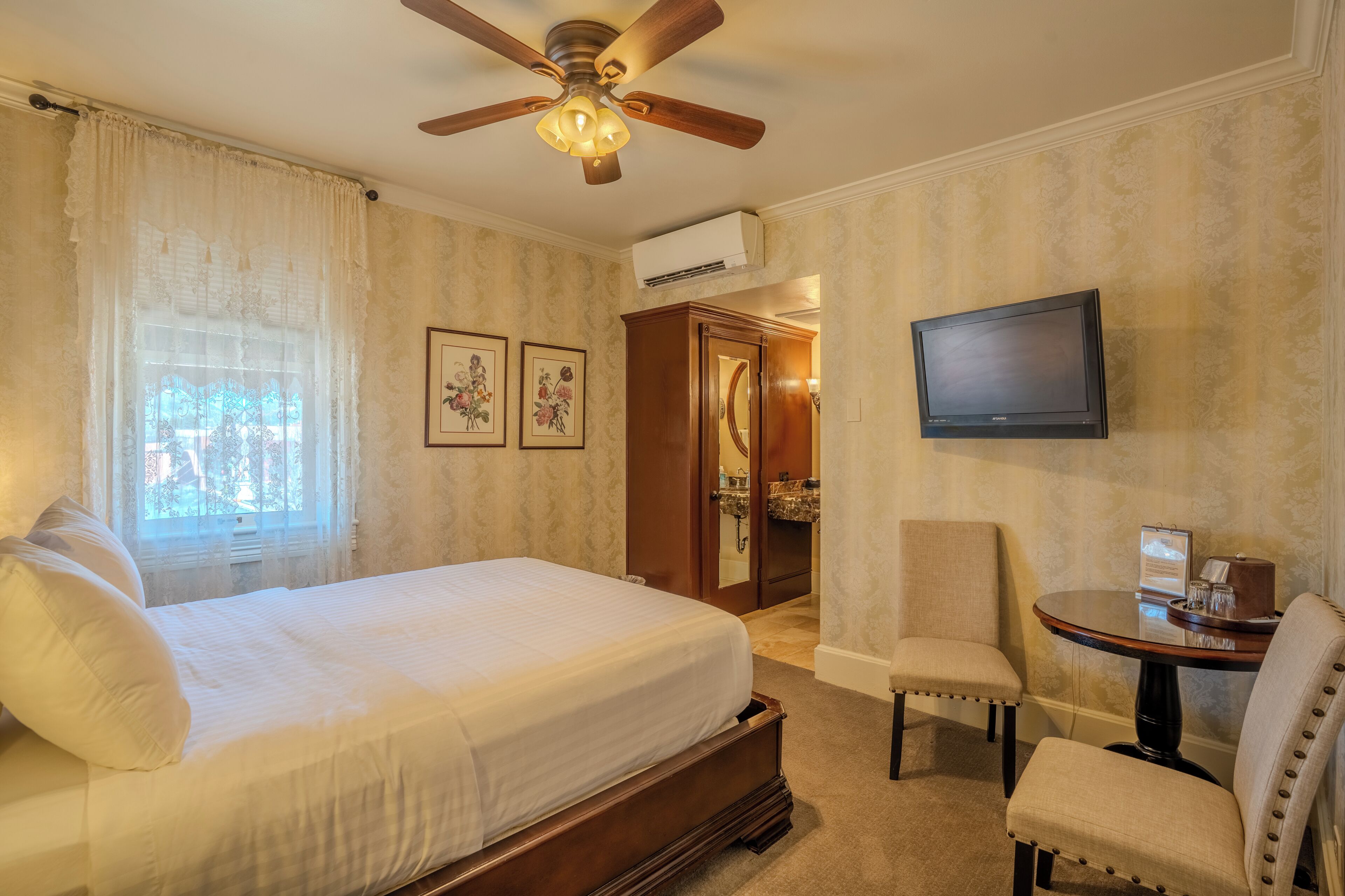 Habitación de hotel con cama, ventilador de techo, televisor y una pequeña mesa con sillas.