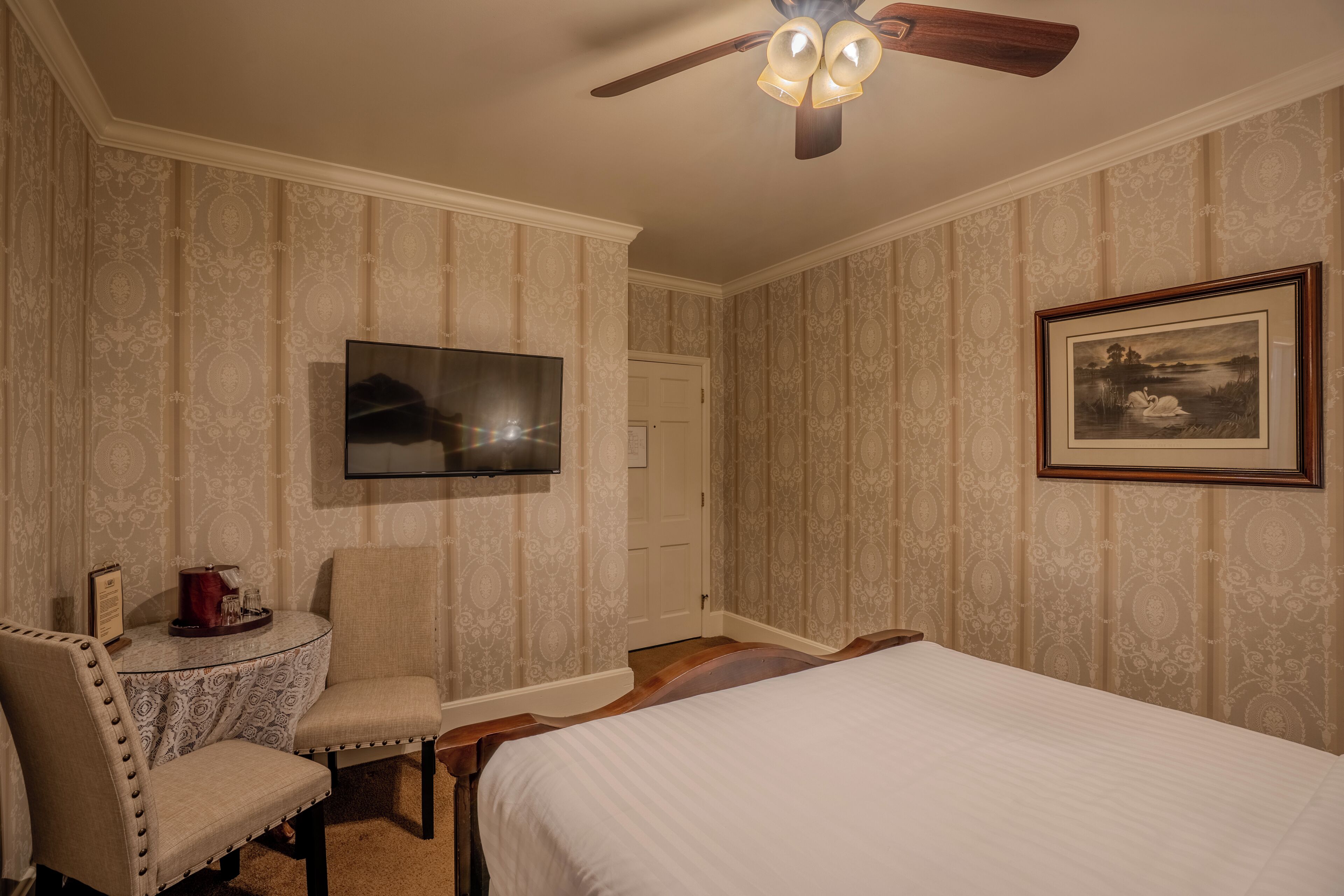 A hotel room with a bed, a chair, a table with a tray, a ceiling fan, a TV, and a framed picture on the wall.