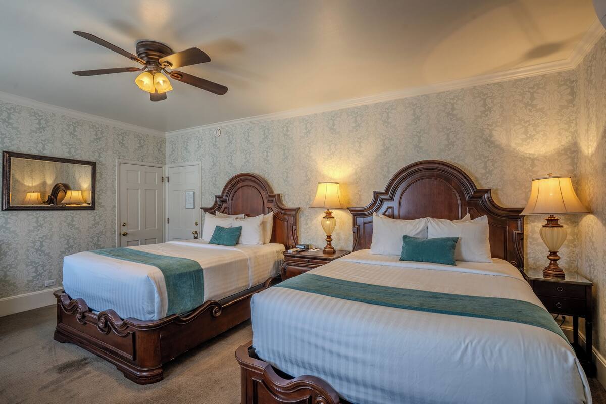 A hotel room with two beds, a ceiling fan, lamps, a mirror, and a door.