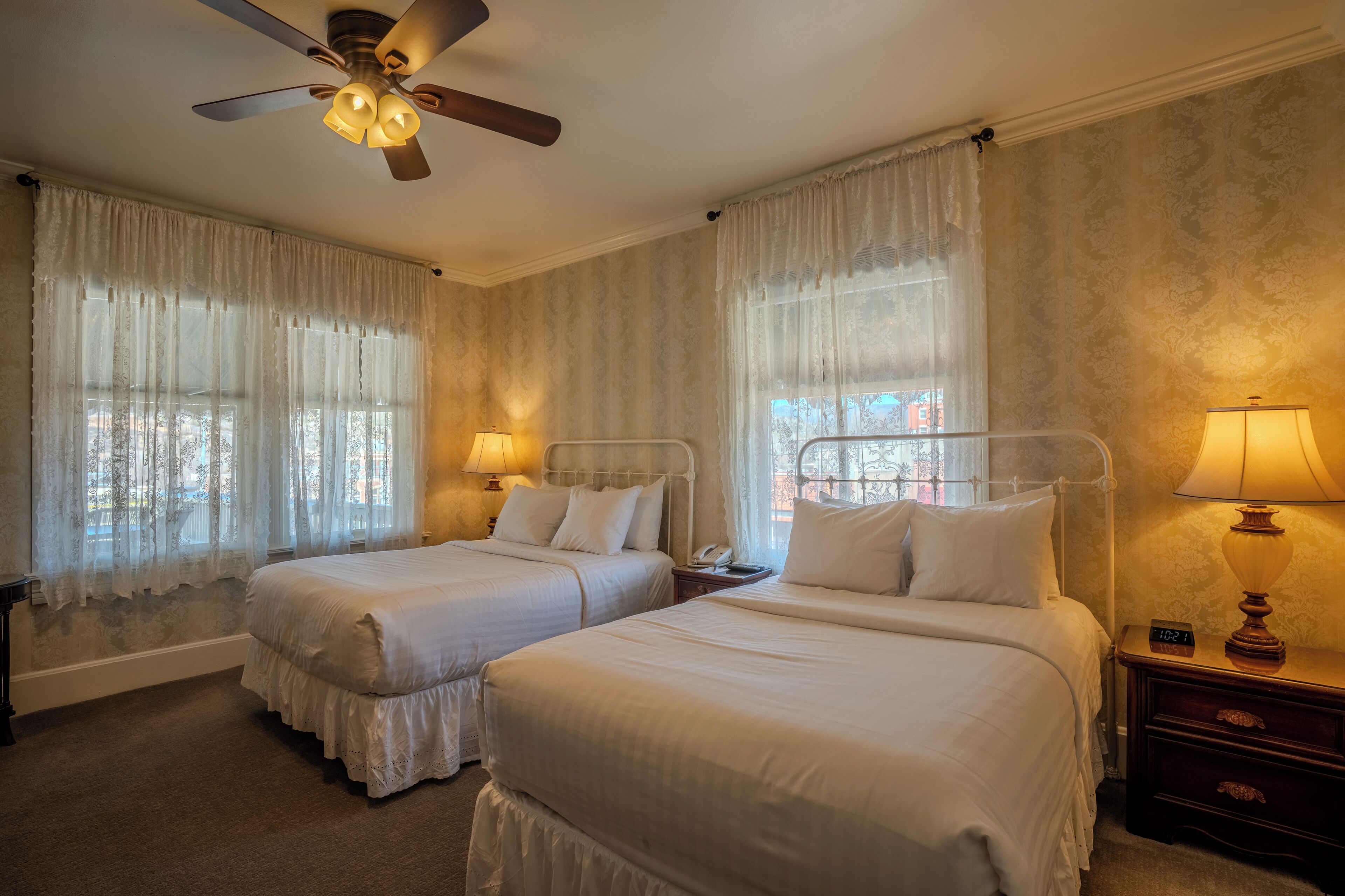 A hotel room with two beds, a ceiling fan, a nightstand with a lamp, and a window with curtains.