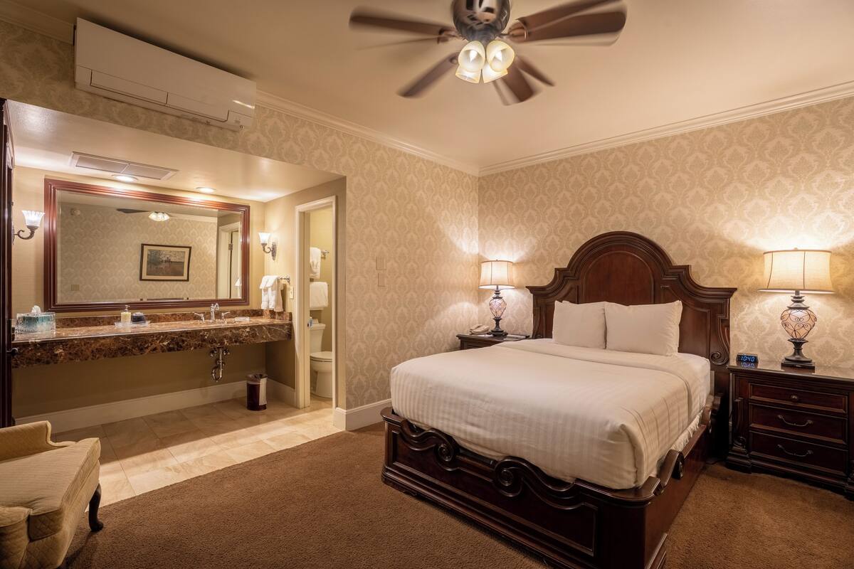A hotel room with a large bed, a vanity with a mirror, a ceiling fan, and a nightstand with a lamp.