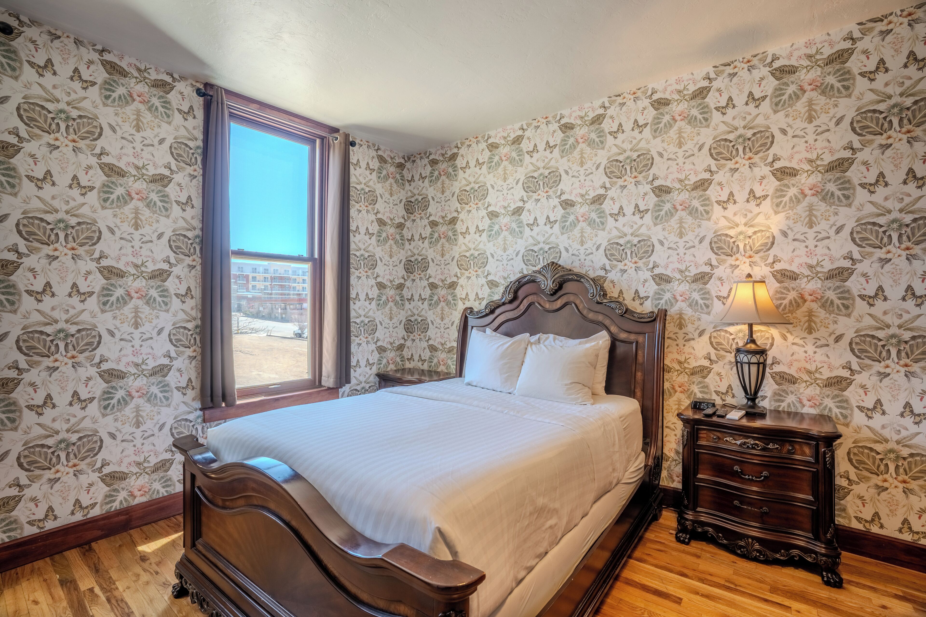 A bedroom with a large bed, a nightstand with a lamp, floral wallpaper, and a window with curtains.