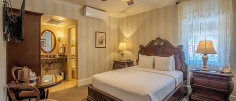 Une chambre à coucher comprenant un lit, un ventilateur de plafond, une table de chevet, une lampe, une chaise, une commode, un miroir et une salle de bain visible à travers une porte ouverte.